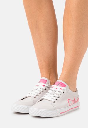 Baskets basses blanches avec des accents roses, des lacets blancs et un logo sur le côté, portées pieds nus sur un fond clair.