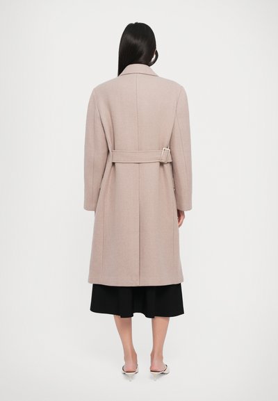 Woman with long dark hair wears beige belted coat, black midi skirt, and white kitten heels, standing against plain white background.