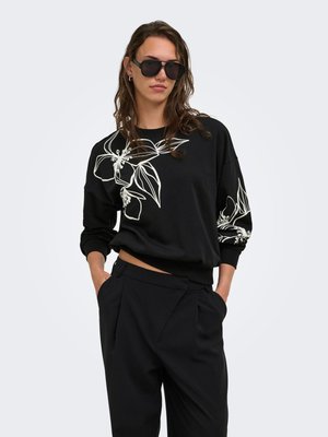 Kvinde iført sorte solbriller, sort sweatshirt med hvidt blomsterstreger, og løstsiddende sorte bukser, står med hænderne i lommerne.
