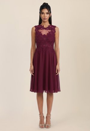 Woman standing wearing a sleeveless knee-length dark red dress with floral lace bodice and sheer neckline, paired with open-toe heels.