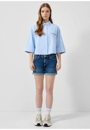 Junge Frau steht, trägt ein hellblau gestreiftes Hemd mit Knöpfen, hochgekrempelte Jeansshorts, weiße Socken und beige Sneakers vor schlichtem Hintergrund.