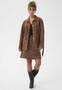 Giacca in suede marrone con dettagli in frange, bottoni dorati e tasche frontali, abbinata a una gonna in suede marrone e stivali cowboy neri.