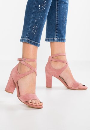 Różowe zamszowe sandały na obcasie z grubym słupkiem, odkryty palec i skrzyżowane paski wokół kostki. Gładka tekstura i minimalistyczny design.
