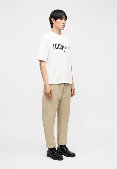 Άνδρας που φοράει λευκό μπλουζάκι με το λογότυπο "ICONique", μπεζ παντελόνι και μαύρα μποτάκια με κορδόνια, στέκεται μπροστά σε απλό φωτεινό φόντο.