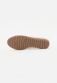 Semelle de chaussure beige avec des rainures horizontales profondes et une zone de talon texturée sur un fond blanc uni.