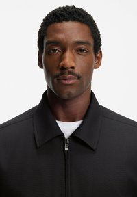 Svart jacka med en strukturerad krage och dragkedja framtill, tillverkad av ett texturerat tyg, kombinerad med en vit t-shirt med rund hals under.