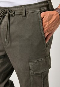 Cargo kalhoty v olivově zelené barvě, vyrobené z odolné tkaniny. Mají elastický pas, šňůrku, boční kapsy a nášivkovou kapsu na noze.