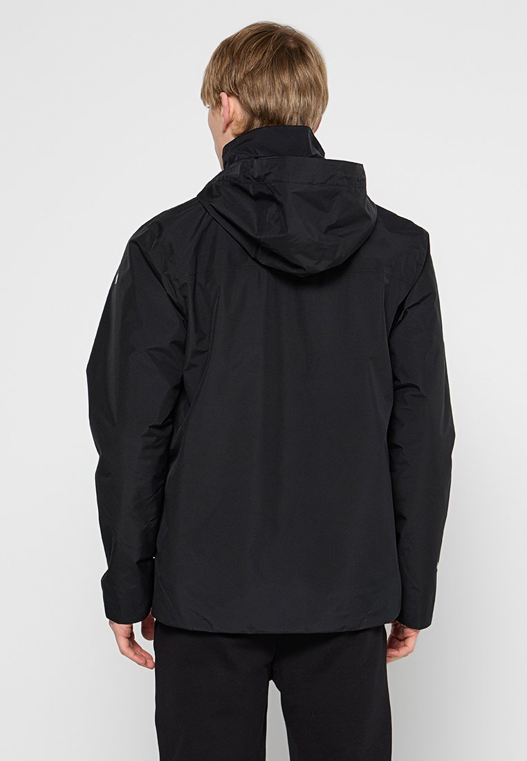 Persoon in een zwarte hoodie en zwarte broek, gezien van achteren tegen een lichte achtergrond.