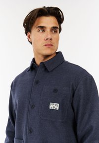 Man in een marineblauwe geknoopte jas met een borstzak en een "Protest"-patch, kijkend naar de zijkant tegen een witte achtergrond.