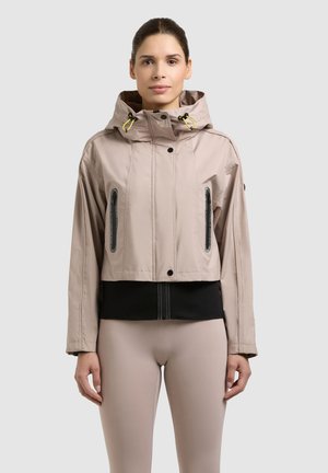 Frau trägt eine kurze beige Kapuzenjacke mit schwarzen Reißverschlusstaschen, steht vor einem schlichten hellgrauen Hintergrund.