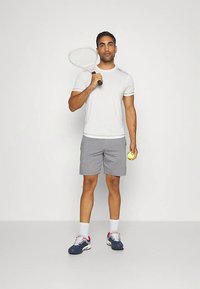 Maglietta sportiva bianca, pantaloncini grigi, tiene una racchetta da tennis in una mano e una pallina da tennis nell'altra, indossa scarpe da ginnastica blu scuro con dettagli rossi.