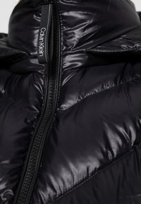 Zwarte donzen jas met een hoge kraag, voorzien van diagonale stiksels en een opvallende ritssluiting. Voorzien van een merkbare trekker voor gemakkelijk gebruik.