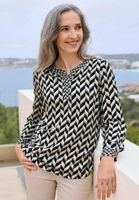 Blouse à motif chevron noir et crème avec un décolleté en V et un détail plissé, associée à un pantalon clair. Tissu lisse et manches longues.