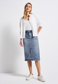 Jupe midi en denim bleu clair avec fente avant et poches, associée à une chemise blanche à boutons et des baskets blanches. Texture lisse.