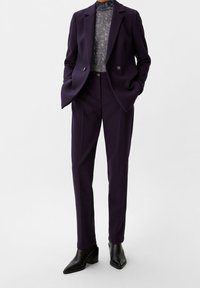 Op maat gemaakt donkerpaars pak met een scherp gesneden jasje en rechte broek. Gecombineerd met een textuur turtleneck en zwarte enkellaarsjes.