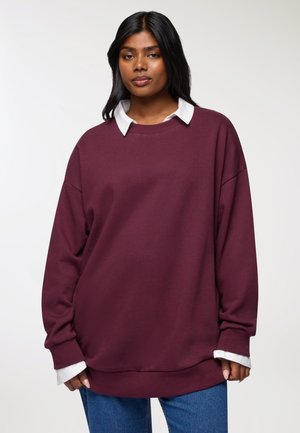 Donna che indossa una felpa over size bordeaux sopra una camicia bianca con colletto, abbinata a jeans blu, in piedi contro uno sfondo chiaro e uniforme.