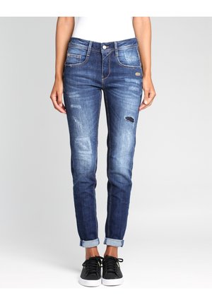 Jeans de mezclilla azul con un corte slim, detalles desgastados y puños enrollados. Presenta bolsillos frontales y costuras doradas. Combinados con zapatillas negras.