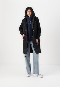 Svart lang frakk med frontglidelås og knapper, kombinert med en marineblå hettegenser og lyseblå ripped jeans. Har quiltsøm nederst.