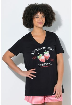 Frau mit lockigem Haar trägt ein schwarzes V-Ausschnitt-T-Shirt mit Erdbeergrafik und dem Text „Strawberry Festival“ und rote gestreifte Shorts, lächelnd.