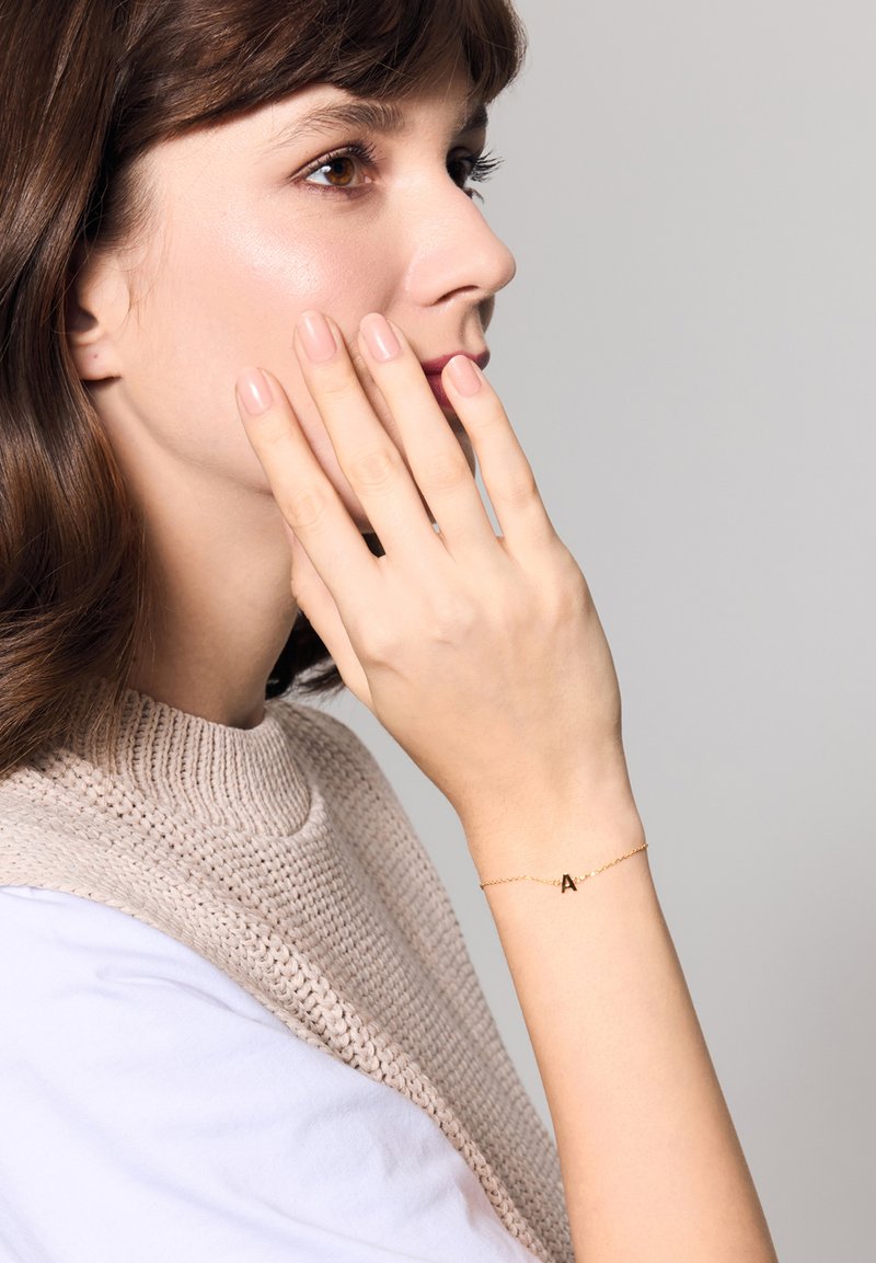 Złota bransoletka z delikatnym łańcuszkiem i zawieszką w kształcie litery "A", noszona na nadgarstku. Modelka ma naturalnie jasne paznokcie i miękki, dziany top.