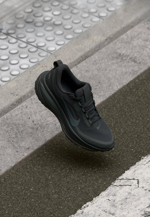 Chaussure de course noire Nike flottant au-dessus d'un trottoir avec un revêtement texturé et des marquages au sol visibles.