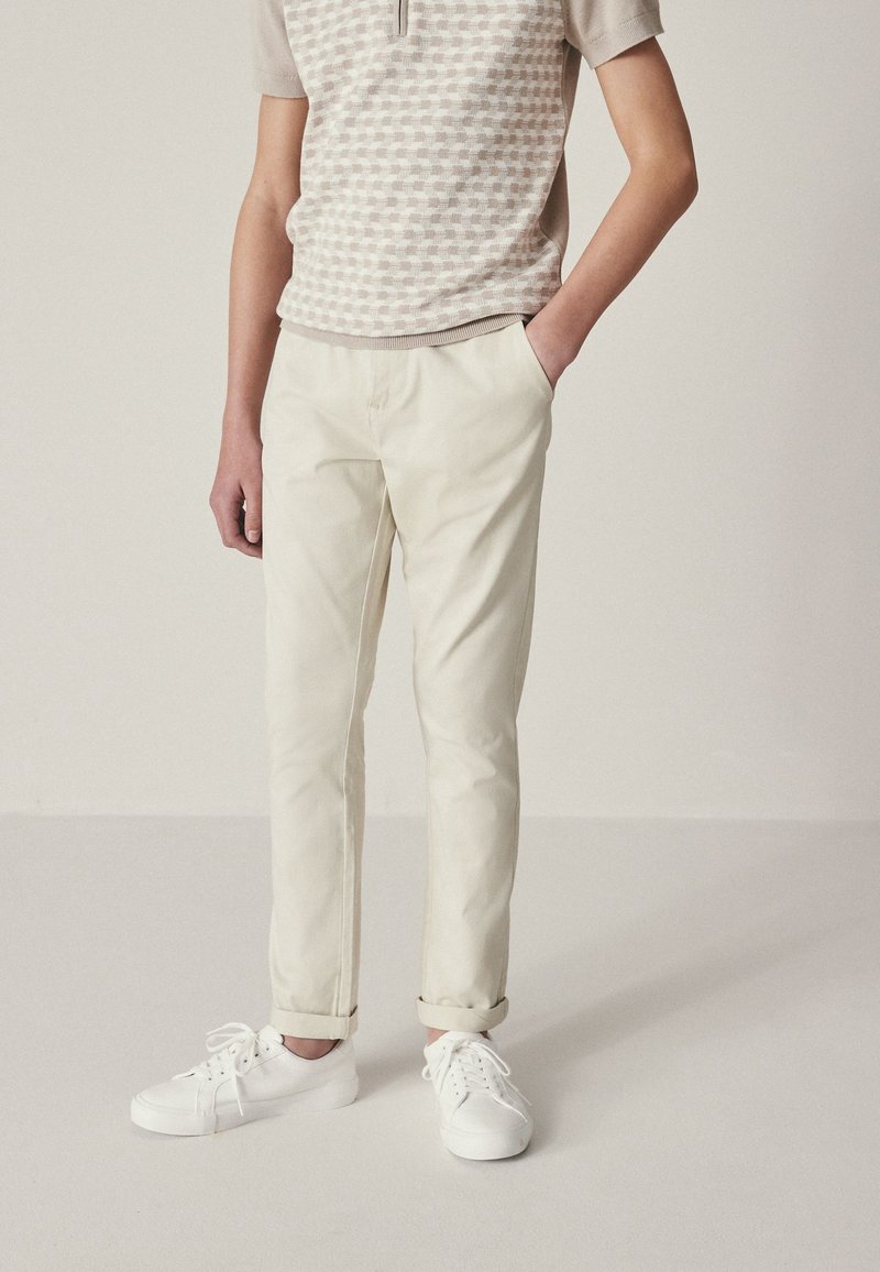 Personne portant une chemise beige à carreaux à manches courtes, un pantalon beige clair avec les revers retroussés et des baskets blanches, debout avec une main dans la poche.