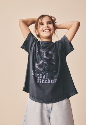 Lächelndes Mädchen mit braunen Haaren in einem schwarzen "Eternal Freedom" Shirt und hellen Jeans hebt die Hände hinter dem Kopf vor einem beigen Hintergrund.