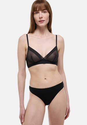 Femme aux cheveux roux lisses portant un bralette noir transparent et un short noir, debout devant un fond blanc uni.