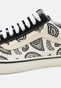 Sneakers with a cream fabric upper featuring a black paisley pattern, black suede accents, white laces, and a white rubber sole with black trim.