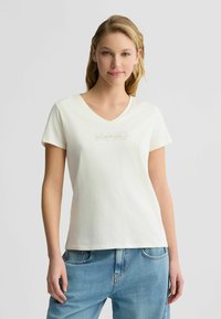 T-shirt V en crème en coton doux avec le mot "iconique" brodé dans un design subtil sur la poitrine, assorti à un denim bleu.