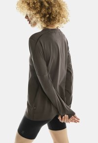 Persoon met krullend haar, gekleed in een donkergrijze lange mouwen shirt en zwarte fietsshorts, staand met de handen gekruist achter hun rug.