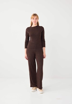 Młoda kobieta stoi nieruchomo, ubrana w pasujący, ciemnobrązowy teksturowany top z długim rękawem i szerokie spodnie, na nogach beżowe trampki, na białym tle.