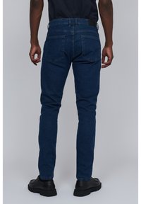 Jeans de mezclilla azul oscuro con un ajuste slim, cinco bolsillos y un acabado texturizado sutil. Presenta un parche en la parte posterior y zapatos negros.