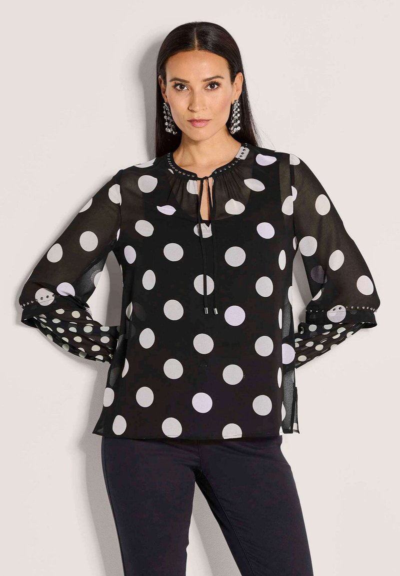 Frau trägt eine schwarze durchsichtige Bluse mit großen weißen Punkten, langen Ärmeln und einer dekorativen Schleife am Hals, kombiniert mit schwarzen Hosen.
