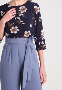 Navy blouse with floral print and 3/4 sleeves paired with high-waisted, light blue trousers featuring a tied waist sash and pleats.