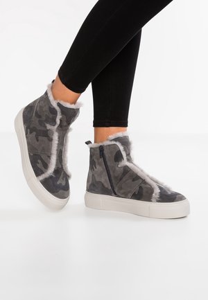 Stopy w szarych, moro wysokich trampkach z białym futrzanym wykończeniem, zamkami błyskawicznymi i rzepami, w połączeniu z czarnymi legginsami.