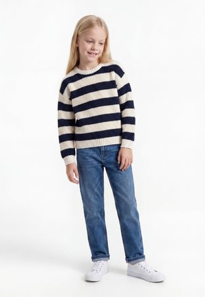 Fille souriante aux longs cheveux blonds, portant un pull rayé noir et blanc, un jean bleu et des baskets blanches, debout sur un fond blanc.