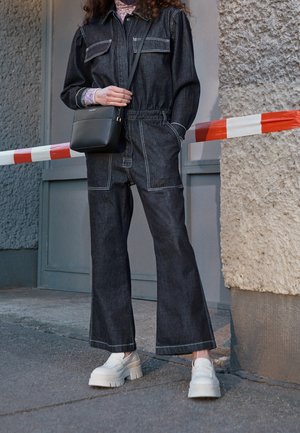 Jumpsuit in denim scuro con maniche lunghe, tasche a toppa e cuciture bianche. Abbinato a scarpe chunky bianche con plateau e una borsa a tracolla nera.
