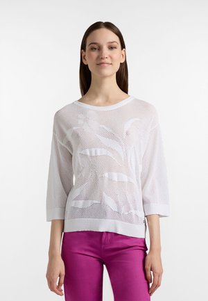 Mladá žena s rovnými hnedými vlasmi oblečená v bielom polopriehľadnom kvetovanom pletenom topu a jasne ružových nohaviciach, stojaca pred bielym pozadím.