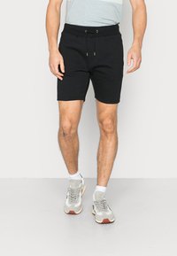 Schwarze Baumwollmix-Shorts mit elastischem Bund, verstellbarem Kordelzug und seitlichen Taschen. Getragen mit hellgrauen Sneakers und weißen Socken.