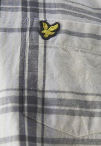 Camisa de cuadros en algodón gris y blanco con un acabado texturizado. Presenta un parche negro con un emblema de águila amarilla en el bolsillo del pecho.