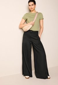 Pantalones anchos negros con pliegues frontales y cortes laterales, combinados con una camiseta verde claro y un bolso cruzado beige.