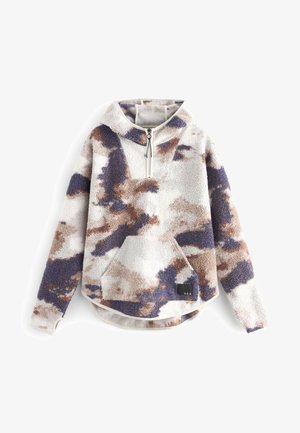 Osoba ubrana w polarowy sweter z fioletowo-brązowymi wzorami, beżowe prążkowane legginsy, białe skarpetki oraz białe buty sportowe New Balance, stojąca na jednolitym tle.