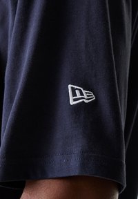 Marineblå t-skjorte med korte ermer og rett nederkant, med en hvit brodert logo på ermene. Laget av myk bomull med en jevn tekstur.