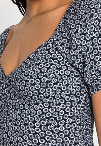 Laivastonsininen mekko, jossa on valkoinen kukkakuosi, lyhyet puff-hihat ja V-mallinen kaula-aukko. Mekossa on kerättyä yksityiskohtaa sivuilla, mikä luo vartalonmyötäisen siluetin.