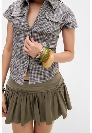 Vrouw die een kortemouwig geruit overhemd en een olijfgroene geplooide rok draagt, houdt de knopen van het overhemd vast met één hand die is versierd met grote armbanden.