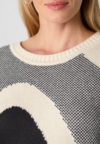 Nahaufnahme einer Frau, die einen schwarz-weißen gestrickten Pullover mit großem Herzmuster trägt, wobei der Ausschnitt und ein Teil des Gesichts zu sehen sind.