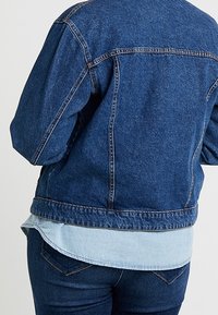 Chaqueta de mezclilla en azul oscuro, con un corte relajado, dobladillo en un azul claro contrastante, y costuras detalladas en los hombros y la espalda.