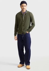 Olivovo zelený pletený kardigán s golierom, zapínaný na gombíky a so žebrovanými manžetami, skombinovaný s tmavomodrými voľnými džínsami a béžovými pracovnými topánkami.