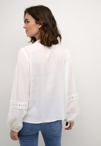 White blouse with long sleeves, featuring puffed cuffs adorned with embroidered accents. Light, sheer fabric with a loose fit design.